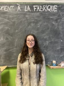 Lilou, 20 ans, jeune femme participante à la performance d'éloquence organisée par l'association Place eux jeunes à Nantes, elle pose devant un tableau noir d'école à la craie et a les cheveux bouclés noirs, elle porte des lunettes noires et un blouse en cuir couleur vert d'eau
