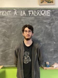 Enes, 18 ans, un jeune participant à la performance d'éloquence de place aux jeunes pose devant un tableau d'école à la craie noir et porte un t-shirt et un gilet gris