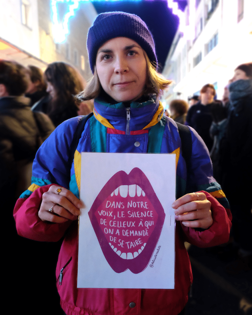 Loreena, membre d'un regroupement de chorales féministes, brandit une illustration de @forme de lutte. Elle représente une bouche féminine ouverte, à l'intérieur de laquelle on peut lire "Dans notre voix, le silence de celleux à qui on a demandé de se taire".Avec la Co chorale, Loreena a interprété plusieurs chants avant les prises de paroles, dont "Cancion sin miedo", de la mexicaine Vivir Quintana, devenu hymne féministe dans toute l'Amérique latine. 25/11/2025 © Amandine Masson