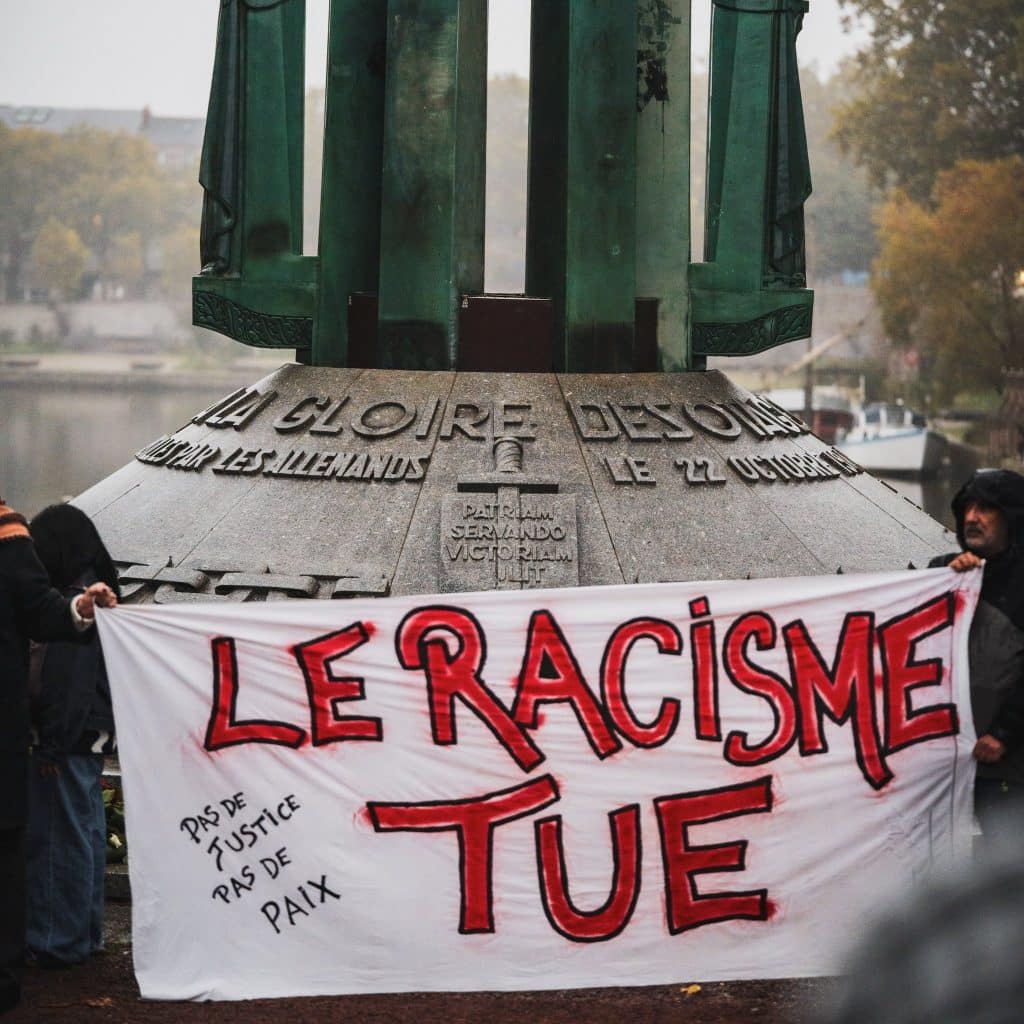 Au pied du monument des 50 Otages et de ses statues vertes, des manifestant·es tiennent une banderole blanche où l'on lit en rouge « Le racisme tue » et, plus petit, « Pas de justice, pas de paix ».