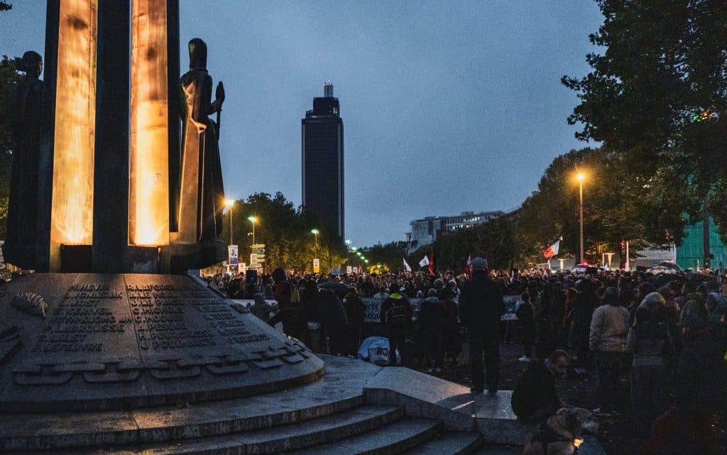 À la tombée de la nuit, le monument des 50 Otages, ses colonnes éclairées et ses noms gravés au premier plan. À droite, la foule rassemblée avec des drapeaux, et la tour Bretagne en fond.