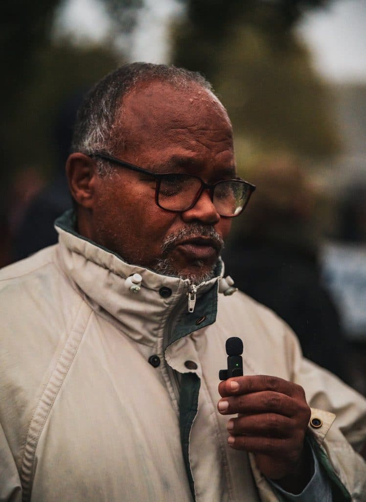 Un homme aux cheveux et à la barbe grisonnants, lunettes et veste beige au col relevé, parle en tenant un petit micro dans la main. Le fond est flou.